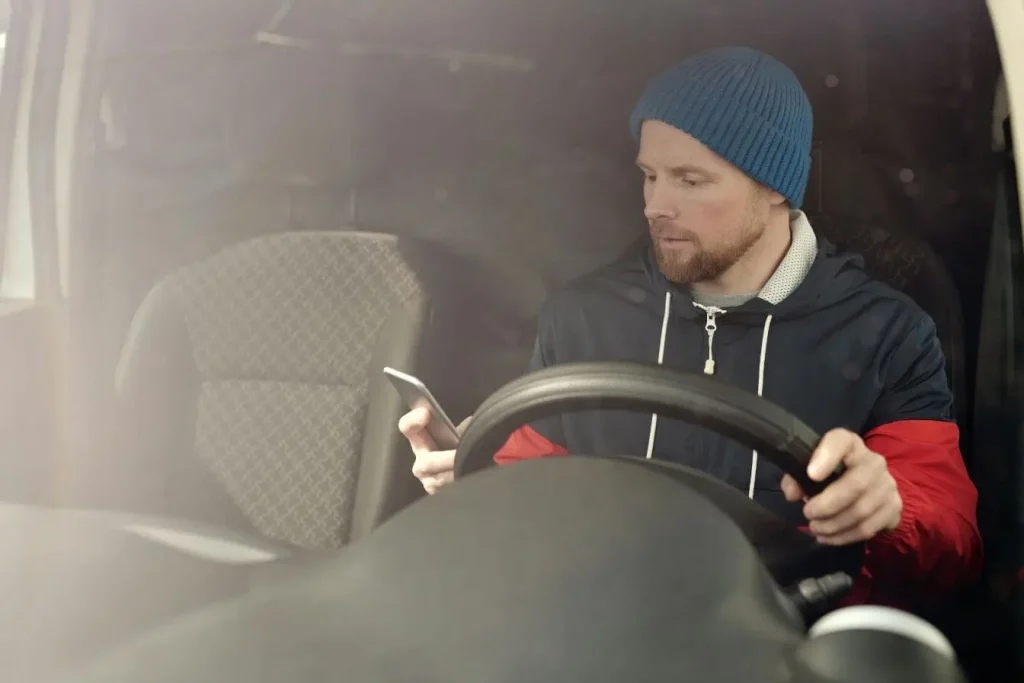 Conducteur portant bonnet bleu tenant smartphone en conduisant, image illustrant le téléphone au volant et les sanctions avec retrait de points en 2026.