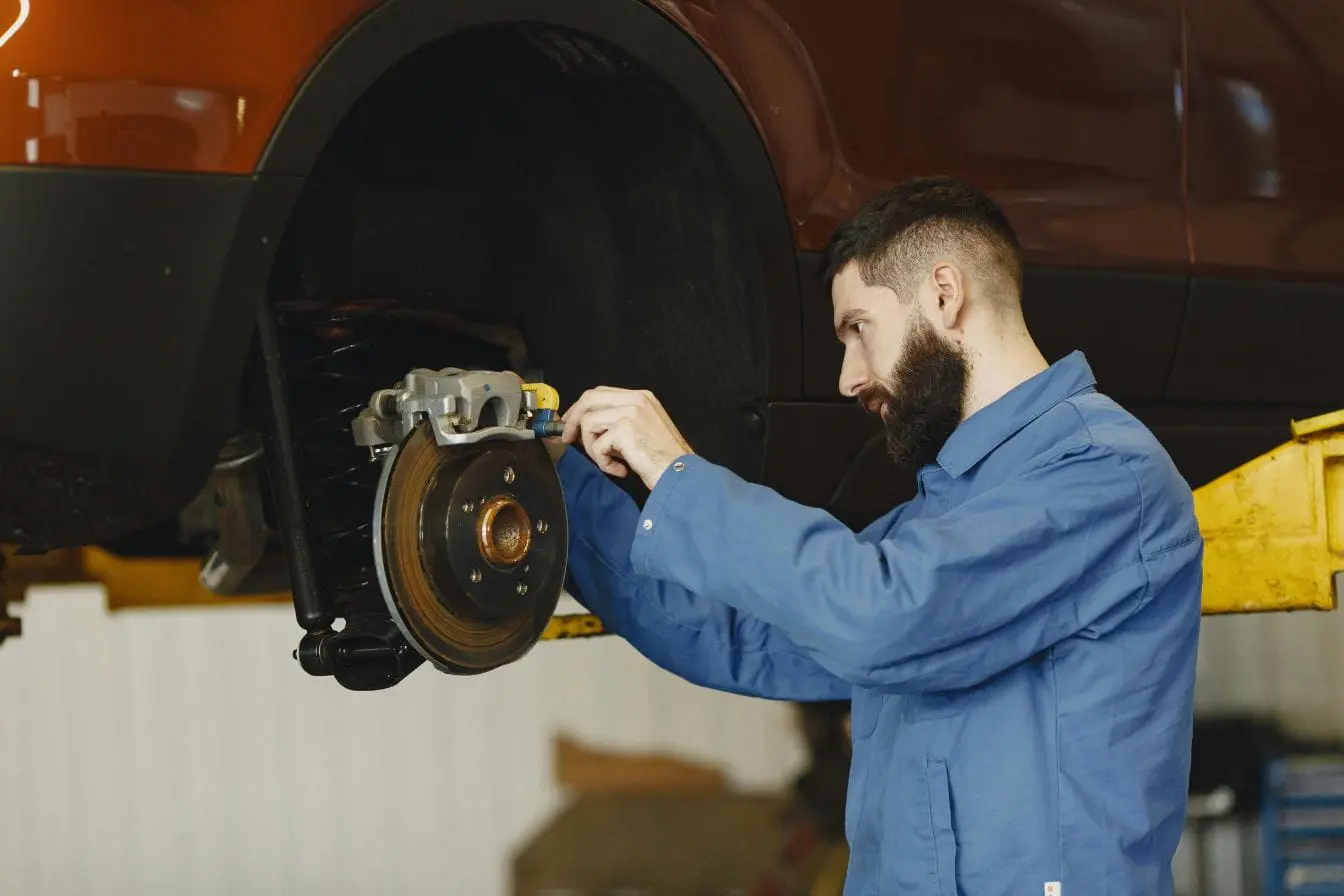 Mécanicien en bleu ajustant étrier de frein sur voiture levée, image illustrant le changement des plaquettes de frein pour assurer sécurité routière.
