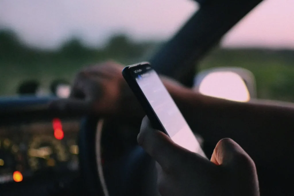 Conducteur tenant smartphone et volant, tableau de bord allumé, image illustrant le téléphone au volant et les sanctions avec retrait de points en 2026.