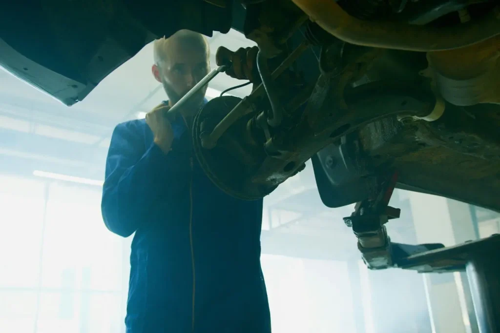 Mécanicien en bleu travaillant sous véhicule levé, image illustrant la réparation d’un cardan qui claque.