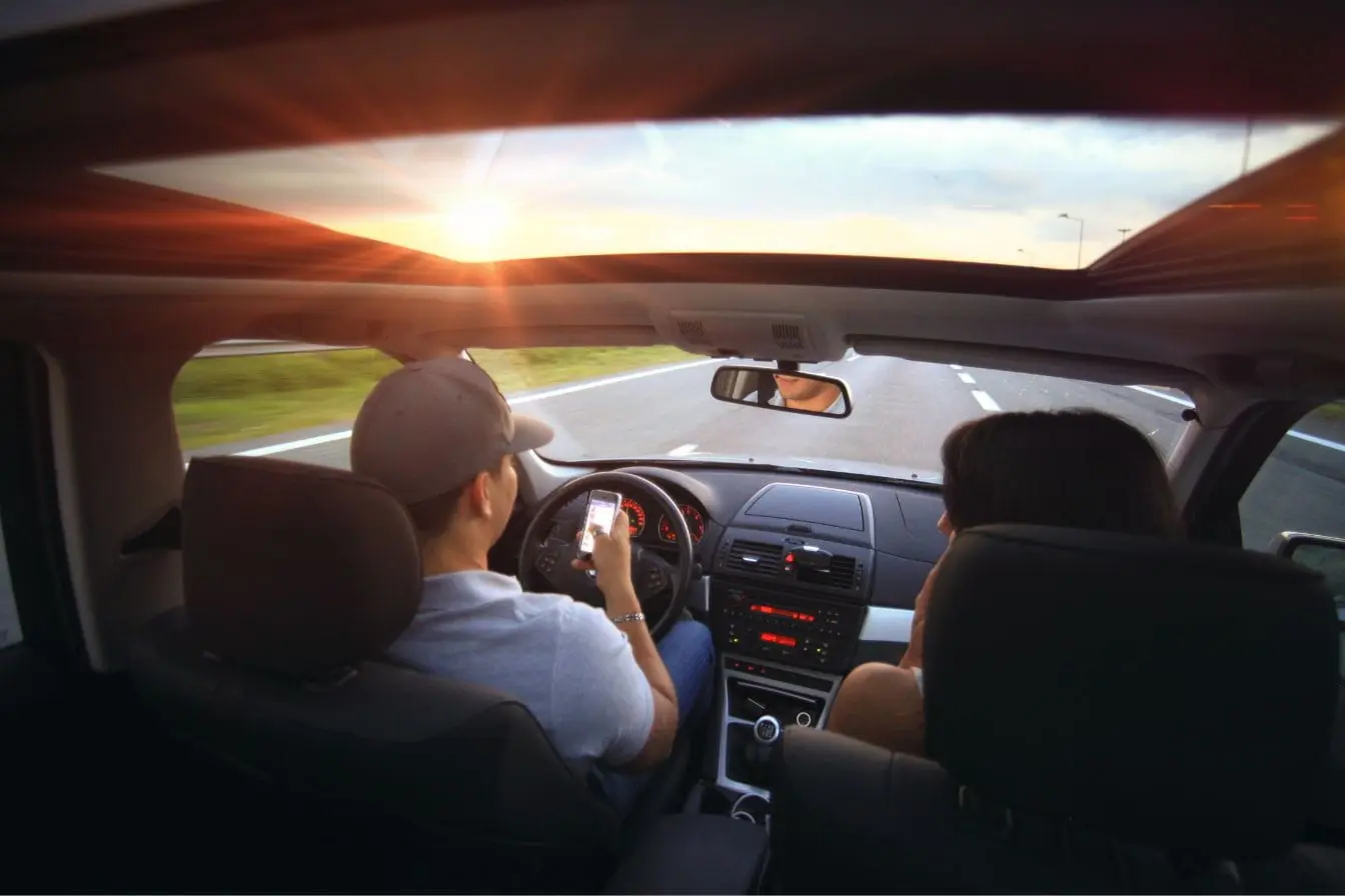 Vue intérieure d’une voiture au coucher du soleil avec conducteur tenant un smartphone, image qui illustre l’importance du système de surveillance des angles morts.