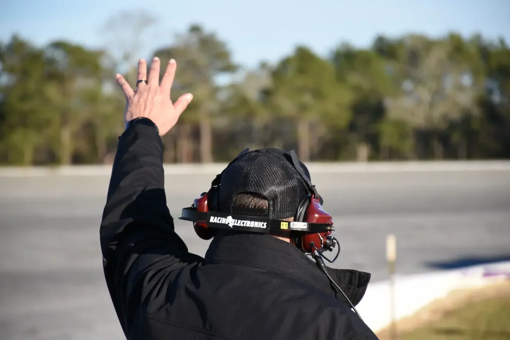 Personne en veste noire portant casque filet et casque audio rouge, geste de signalisation sur piste, symbole des missions pour devenir commissaire de course automobile.