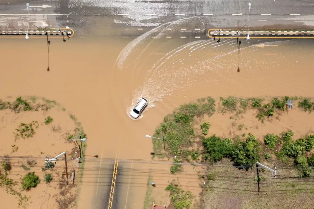 Vue aérienne d’une route inondée avec voiture blanche avançant dans l’eau, reflet des réflexes à adopter après catastrophe naturelle.