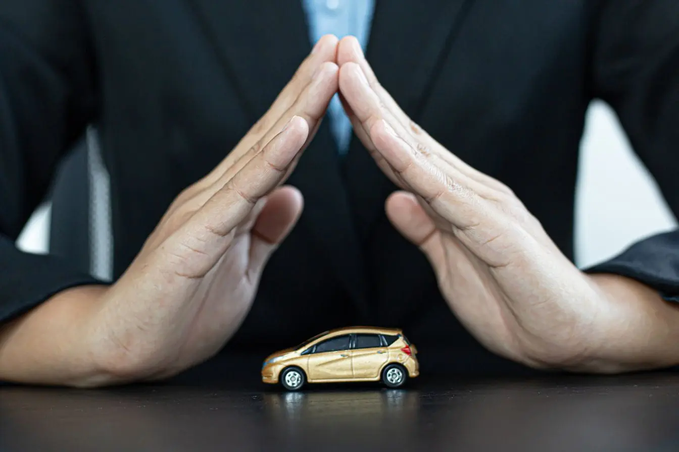 assurance auto Personne en costume protégeant une petite voiture dorée avec ses mains, symbole de l’assurance auto pour véhicule de collection.